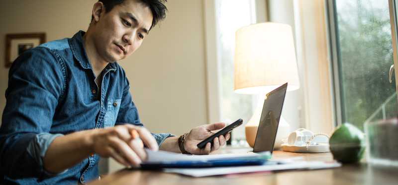 Person sitting at computer while reviewing paperwork and holding phone.