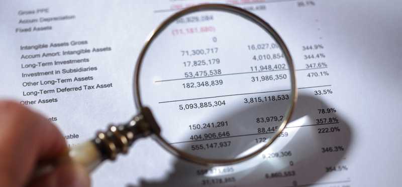 A hand holding a magnifying glass up to some financial paperwork.