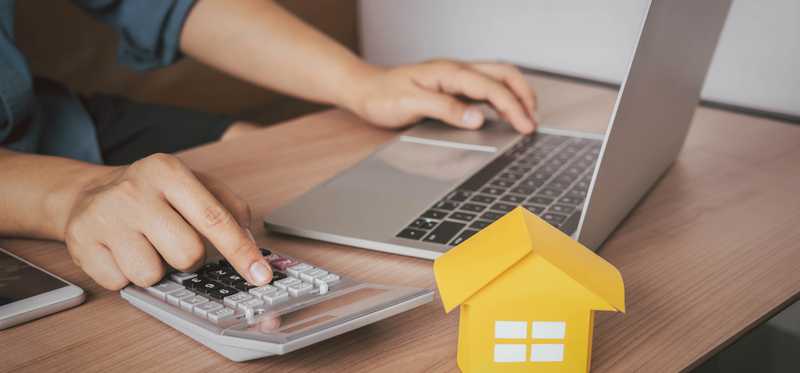 A person working on a laptop and a calculator next to a small model of a home sitting on the desk.