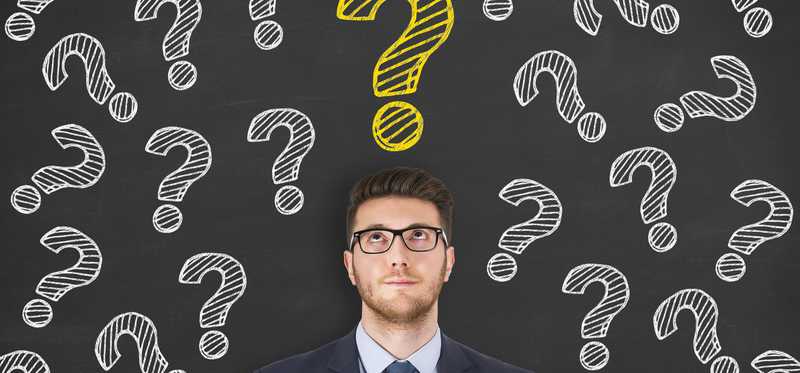 Professionally dressed young man standing in front of chalk board with question marks drawn over his head.