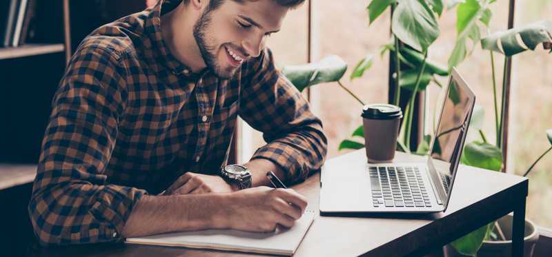 Young man writing in notebook with laptop open in front of him.