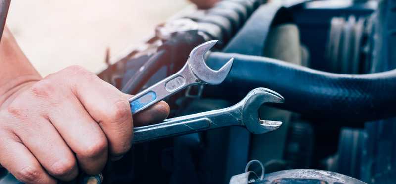 Mechanic holding wrenches and looking under hood of car.