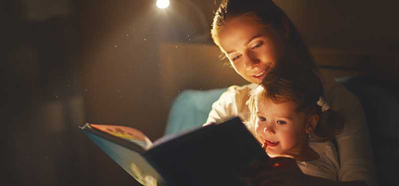 Mother reading book to young daughter in dark room.