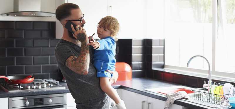 Adult holding child and talking on phone in kitchen.