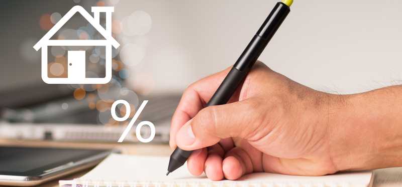 A hand with a pen writes in a notebook with a percent sign and the outline of a house hovering over it.
