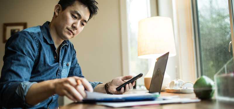 Person holding smartphone and looking at document in home office.