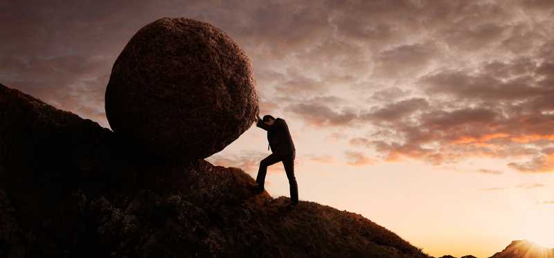 Man pushes a boulder up a hill with sunset behind him.
