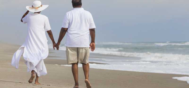 A retired couple walk hand in hand on the beach.