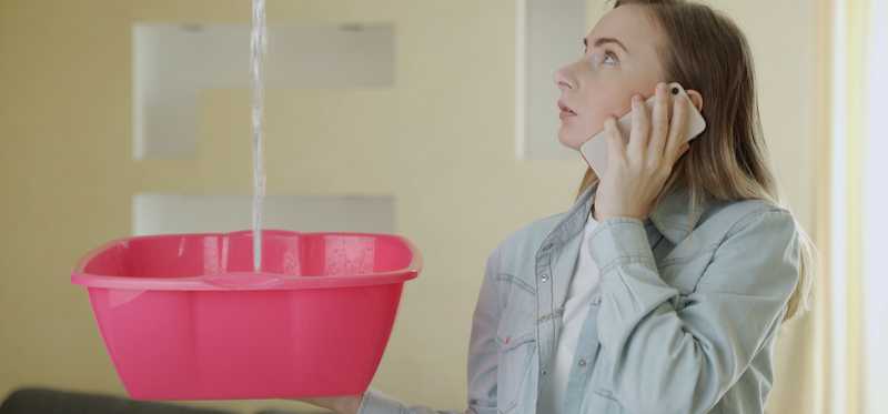A person on the phone while holding a bucket to catch water leaking from the roof.