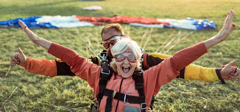 Two happy people in skydiving gear with parachute and plane behind them.