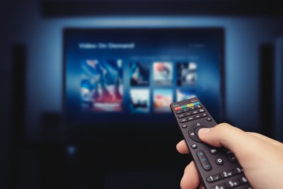 Person holding remote against background of TV with streaming service on screen.