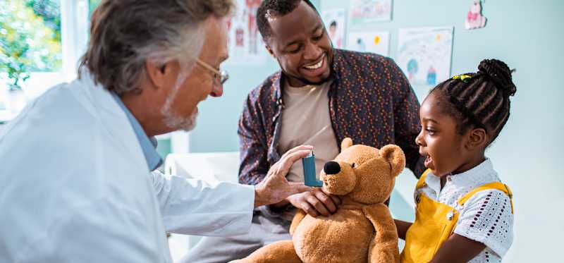 Doctor using a teddy bear to show a girl and her father how inhaler works