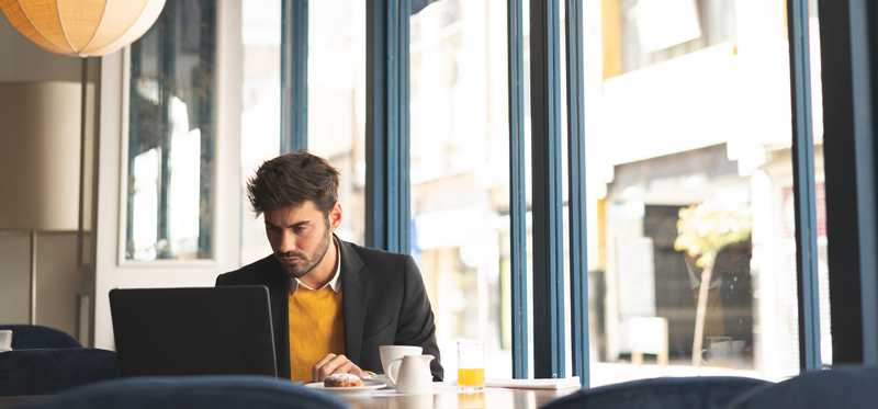 Person sitting with laptop and breakfast items on table by large window with sun pouring in.