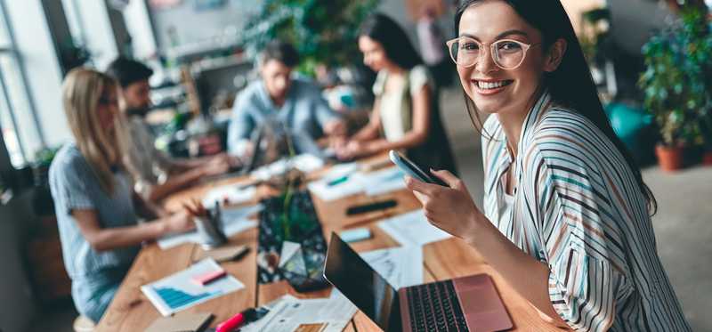 Person in striped shirt and glasses is smiling and sitting at long wooden table with other remote workers.