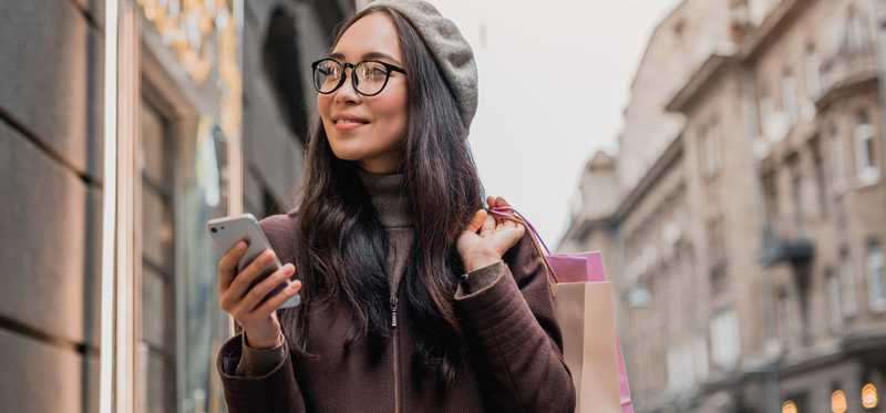 Woman in beret looks at smartphone while walking down street and holding shopping bag
