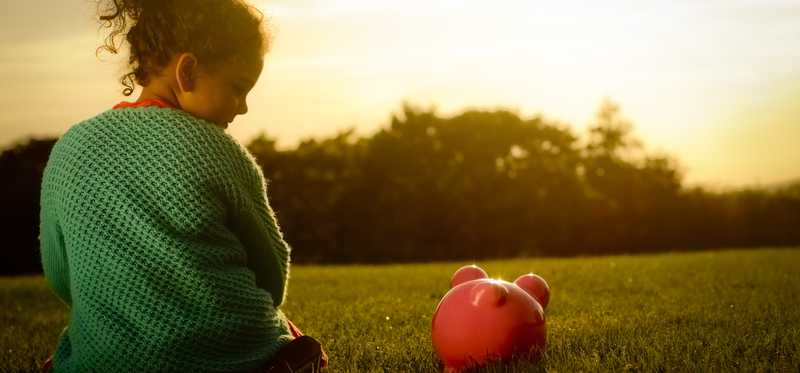 Young girl sitting next to piggy bank on lawn and watching the sun set
