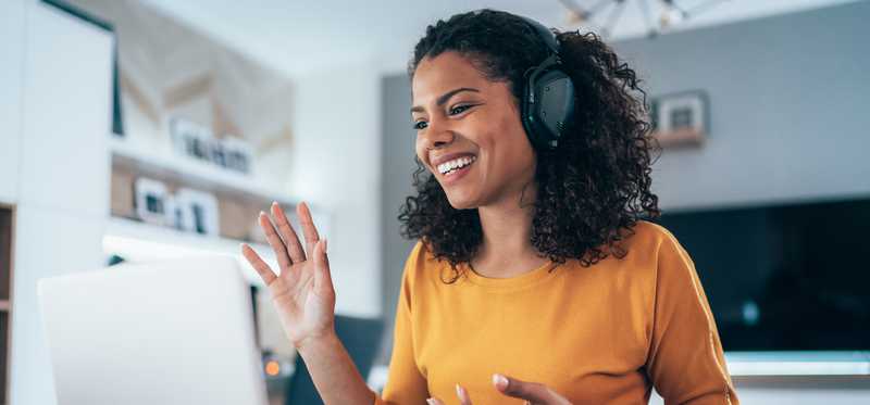 Woman wearing headphones while sitting at kitchen table and waving to coworkers on videoconference