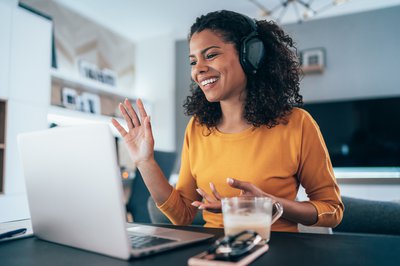 Woman wearing headphones while sitting at kitchen table and waving to coworkers on videoconference