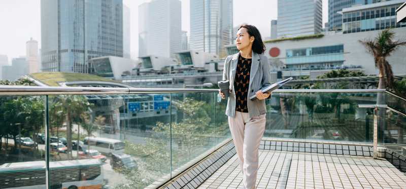Businessperson walking with folder of papers on rooftop balcony and staring out at cityscape.