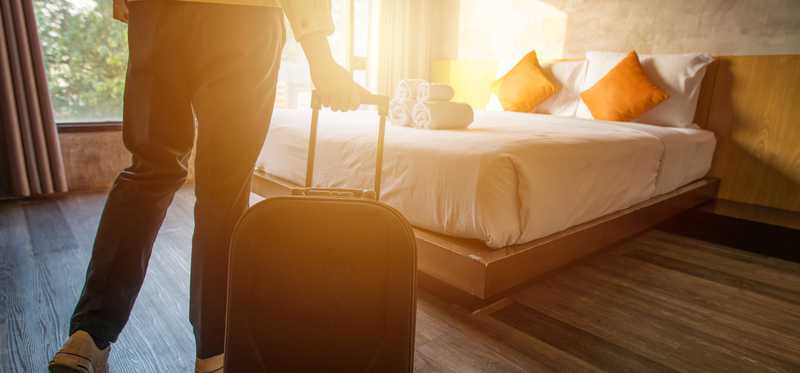 Person rolling carry-on luggage near bed in sun-filled hotel room.