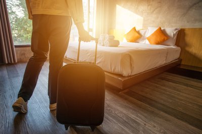 Person rolling carry-on luggage near bed in sun-filled hotel room.