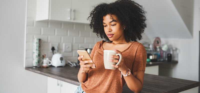 Woman leaning against kitchen countertop while looking at phone and holding cup of coffee