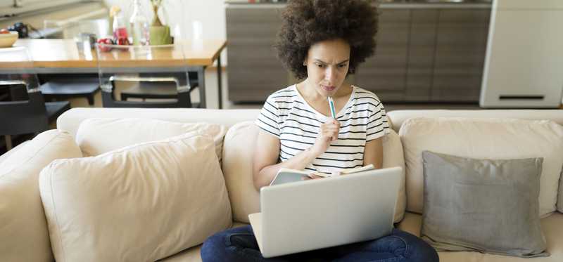 Woman sitting cross-legged on couch while looking at laptop. with perplexed expression