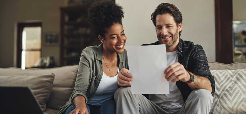 Two people sitting on couch and smiling while looking at paperwork.
