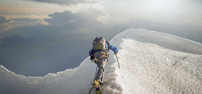 Hiker climbing snowy mountain against cloudy sky.