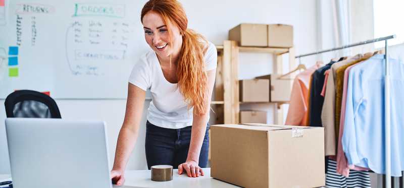 Woman leaning over desk and computer with boxes and clothing rack.