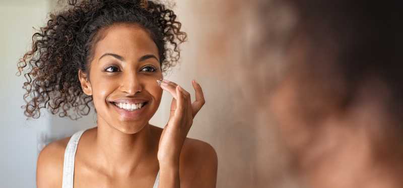Young woman standing in front of mirror applying face cream
