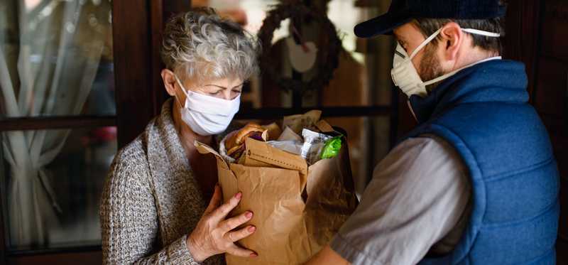 Food delivery worker in mask handing brown paper bag of food to masked person..