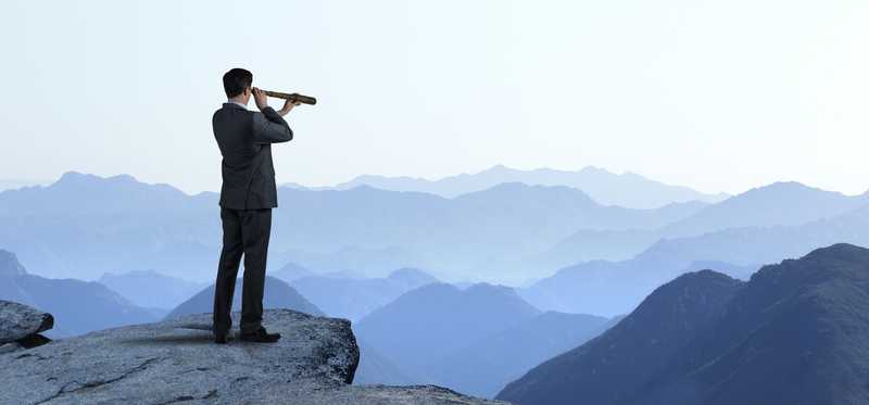 Person standing on cliff is using telescope to look out at mountains on horizon.
