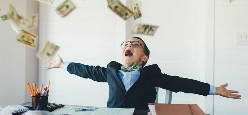 Money raining down on child dressed in suit and sitting at desk.
