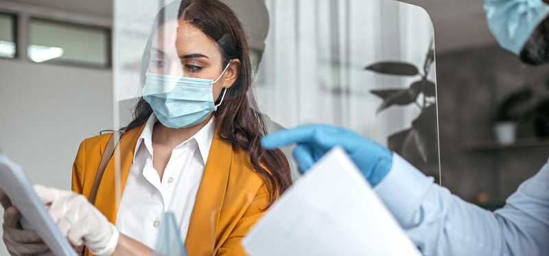 Doctor wearing personal protective equipment speaking through glass barrier to patient wearing mask and gloves