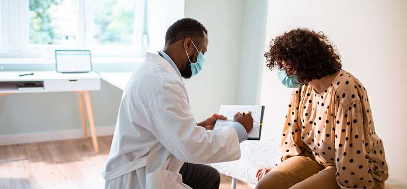 Doctor in mask and lab coat talking with masked patient about test results.