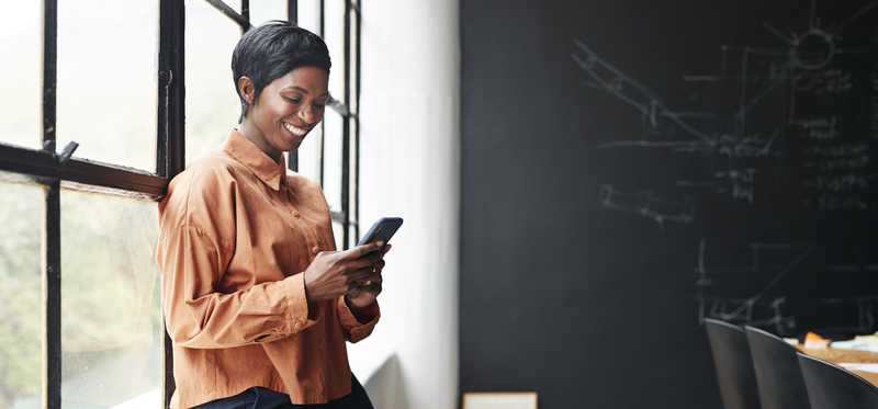 Smiling person holding phone while sitting on windowsill in sunlit room.