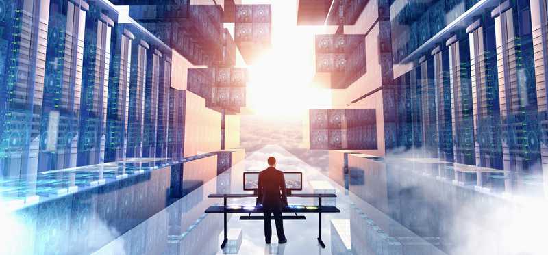 Worker sitting at dual monitor at desk in illuminated server room against backdrop of glowing sky.