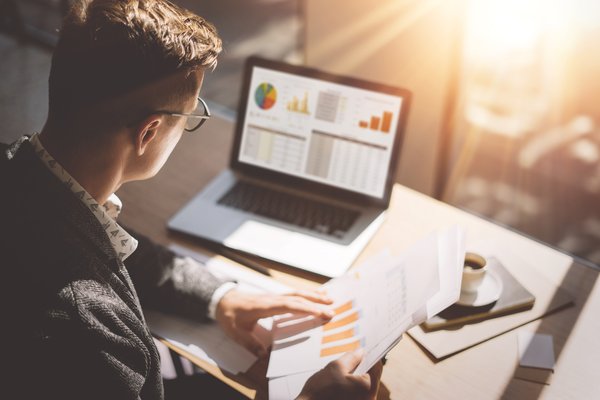 Person looking at graphs on laptop and paperwork.