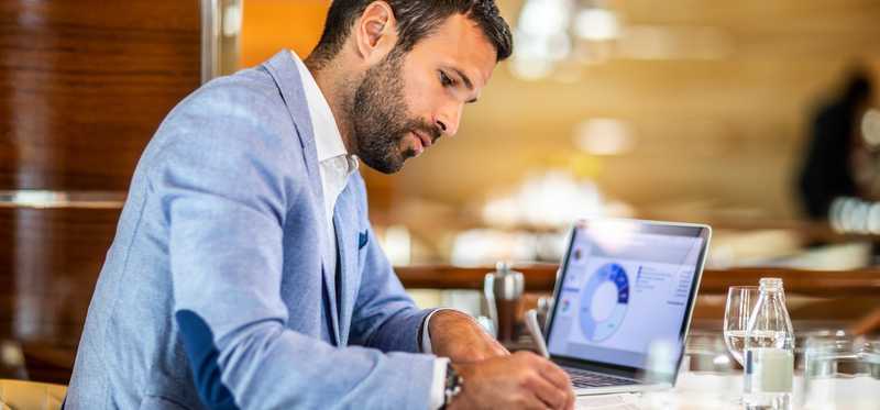 Businessperson reviewing chart on laptop and writing notes.