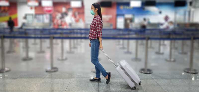 Masked person in checkered shirt walking through mostly empty airport rolling carry-on luggage.