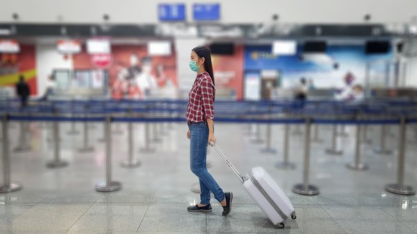 Masked person in checkered shirt walking through mostly empty airport rolling carry-on luggage.