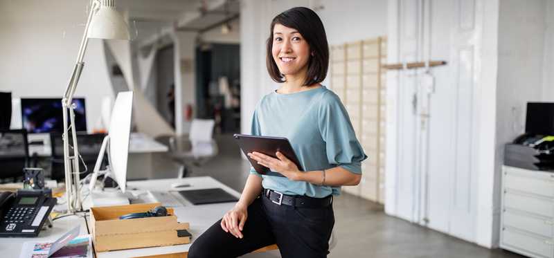 Smiling person holding tablet while standing and leaning on a desk in an office.