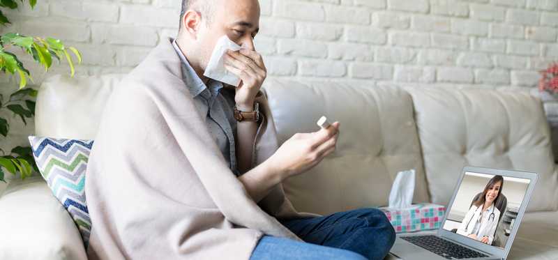 Sick person wrapped in blanket and blowing nose while attending telehealth appointment.