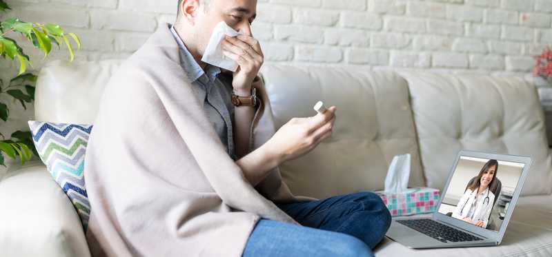 Sick man wrapped in blanket and blowing nose attending telehealth appointment