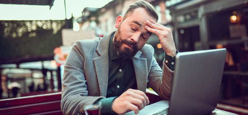 Stressed person with hand to forehead is sitting at outdoor cafe with an open laptop.
