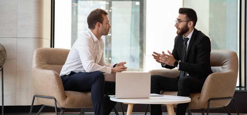 Two businesspeople sitting in tan chairs talking.