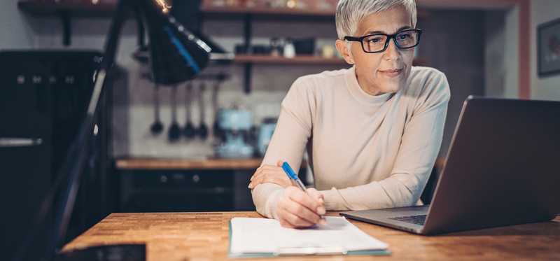 Person sitting at laptop in kitchen and writing on paper.