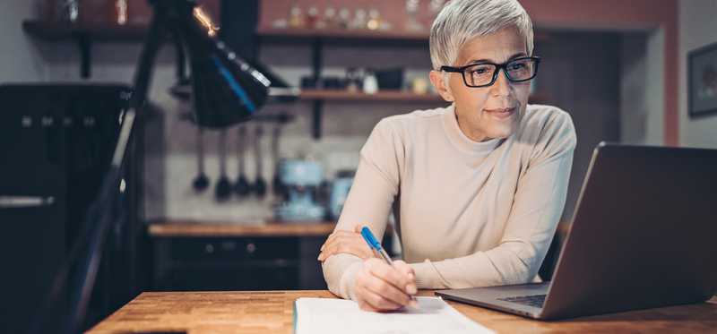 Person sitting at laptop in kitchen and writing on paper.