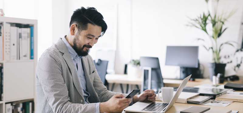 Person sitting at table with laptop while smiling and looking at phone.